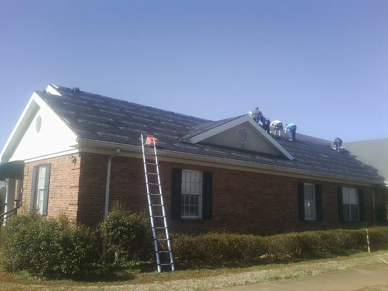 Crew tearing shingles off a brick ranch roof with a ladder set — Metal Roof Repair work by our crew in Ooltewah