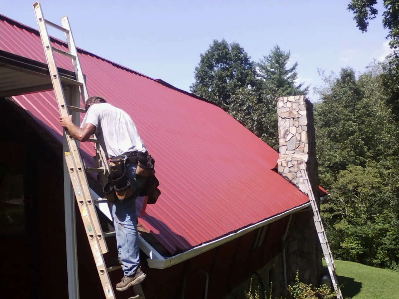 Roofer on a ladder beside a red metal roof and stone chimney — Soffit & Fascia Repair by Roofing Brothers in Ooltewah